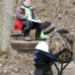 Two children sitting in folding chairs in the woods drawing their surroundings on a notepad.