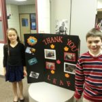 Two children proudly standing in front of a trifold poster board that says "Thank You"
