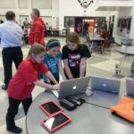 Three young girls hovering over a single laptop surrounded by additional laptops and tablets.