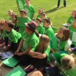 A group of children, all wearing green shirts, sitting on the grass listening to a teacher.