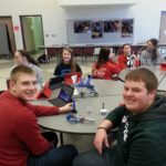 Three students sitting at a cafeteria table with laptops and science materials smiling.