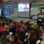 A group of young students sitting in a classroom setting all looking toward a projector screen on the wall in front of them.