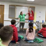 Children sitting on the floor looking at two teachers who are presenting a science experiment using a beaker of liquid.