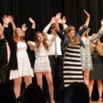 A group of teenage girls, all wearing black, white, and gray, with their arms up in the air as if dancing in a routine.