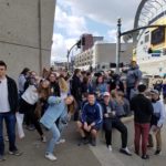 A group of teenage students standing and sitting on a sidewalk across the street from a sports stadium.