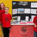 A group of students and a teacher all wearing red shirts standing in front of a poster board reading "Bringing Bladeless Wind Turbines to Small Towns".