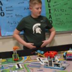 Child in a dark green shirt standing in front of a table filled with Lego robotics.