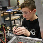 Teenage boy wearing protective eyewear tinkering with a electronic parts in front of him.