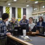 Group of teenage students and a teacher standing around a table in an engineering classroom.