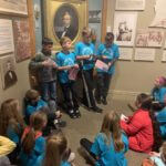 Children wearing blue school shirts standing and sitting in a historical room.
