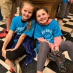 Two young girls sitting on the floor of a museum smiling at the camera.