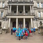 Large group of young students with a teacher standing on the steps of a governement building.