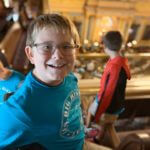 Young boy wearing a blue shirt and glasses smiling at the camera in a historic room.