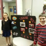 Two young students standing on either side of a black trifold poster board reading "Thank You."