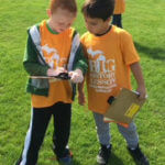 Two young students wearing yellow shirts and holding clipboards working together.