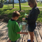 A young student and his teacher carrying a metal bucket outside.