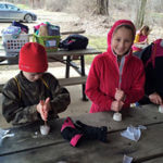 Two children trying to make a fire with a fire starter on a picnic table outside.