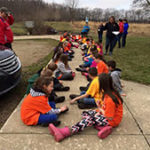 Group of young students sitting down on a sidewalk in two rows facing each other.