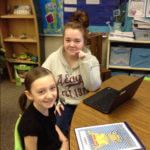 One younger student and one older student sitting at a desk together in front of a chromebook.