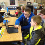 Three students sitting at a desk playing with a chromebook.